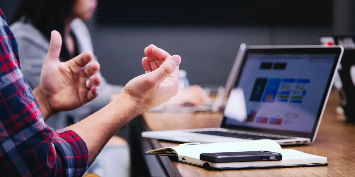 A person gestures during a team meeting at a conference table with a laptop and notebook in front of them.