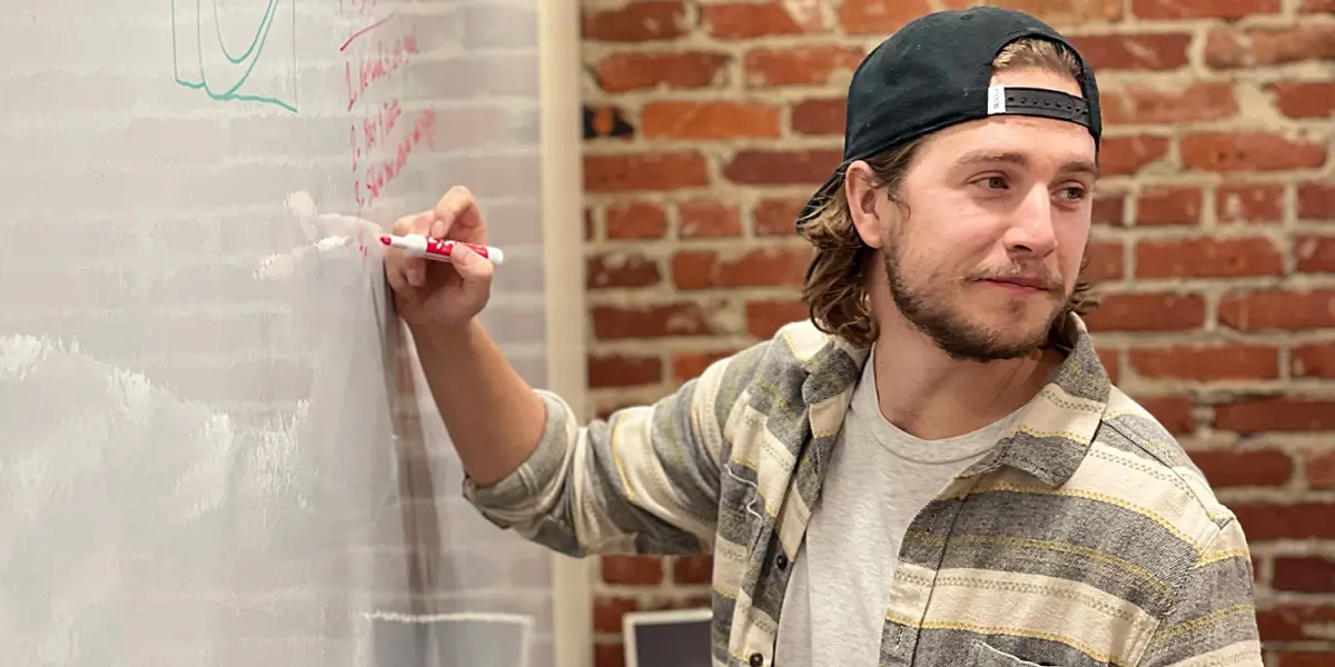 A man in a striped shirt and backwards cap writes on a whiteboard in front of a brick wall during a brainstorming session.