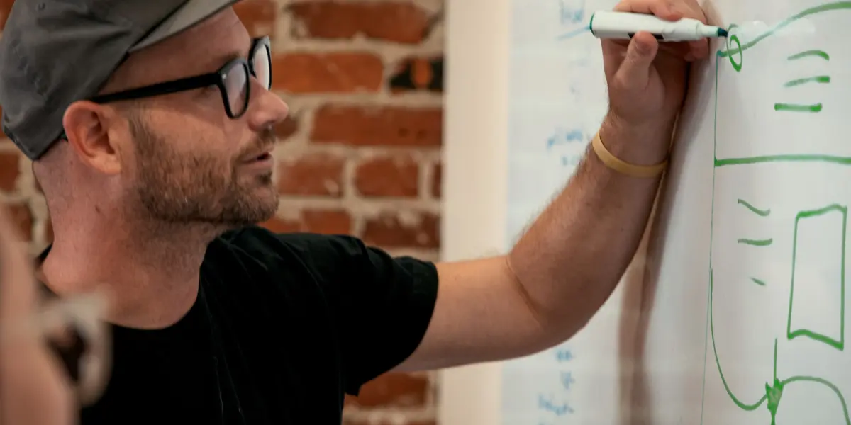 A man in glasses and a cap draws a diagram on a whiteboard during a brainstorming session.
