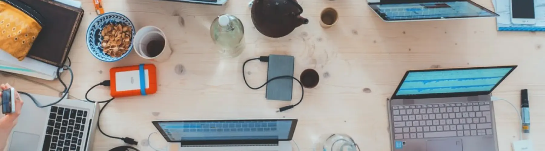A busy shared workspace with multiple laptops, smartphones, notebooks, and snacks spread across a wooden table during a collaborative work session.