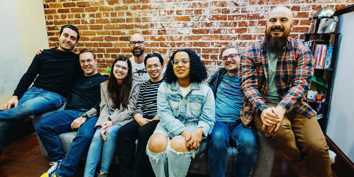 A group of Hounder employees sit together on a couch in front of a brick wall, smiling and relaxed.