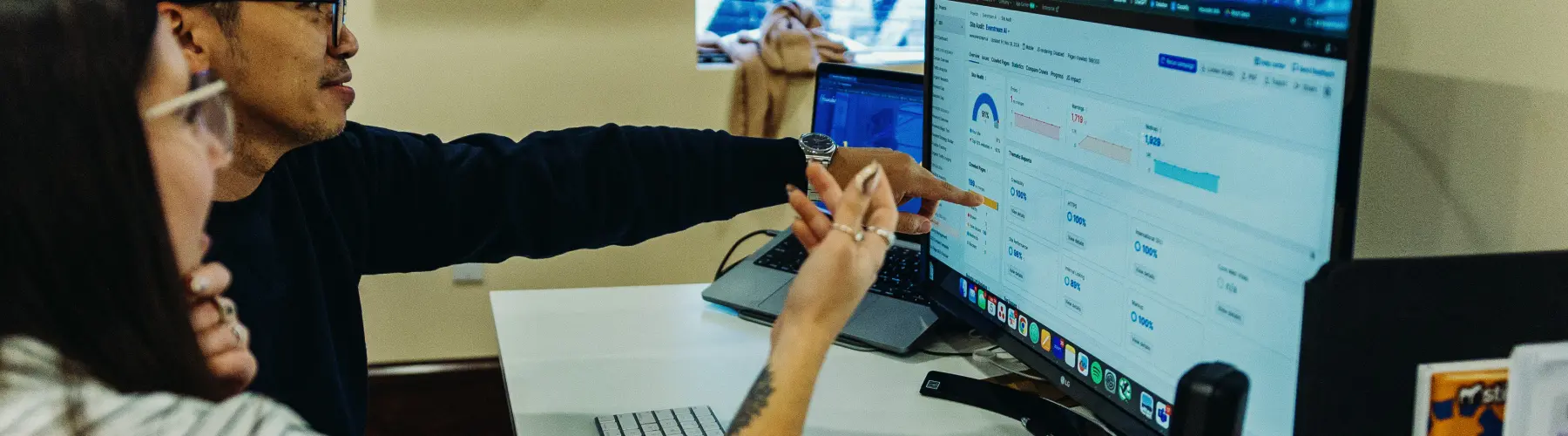 Colleagues review analytics data on a desktop monitor, pointing at various metrics during a discussion.