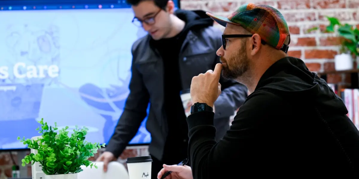 Two Hounder team members collaborate in a modern brick-walled workspace, with one seated and thinking while the other prepares to present.