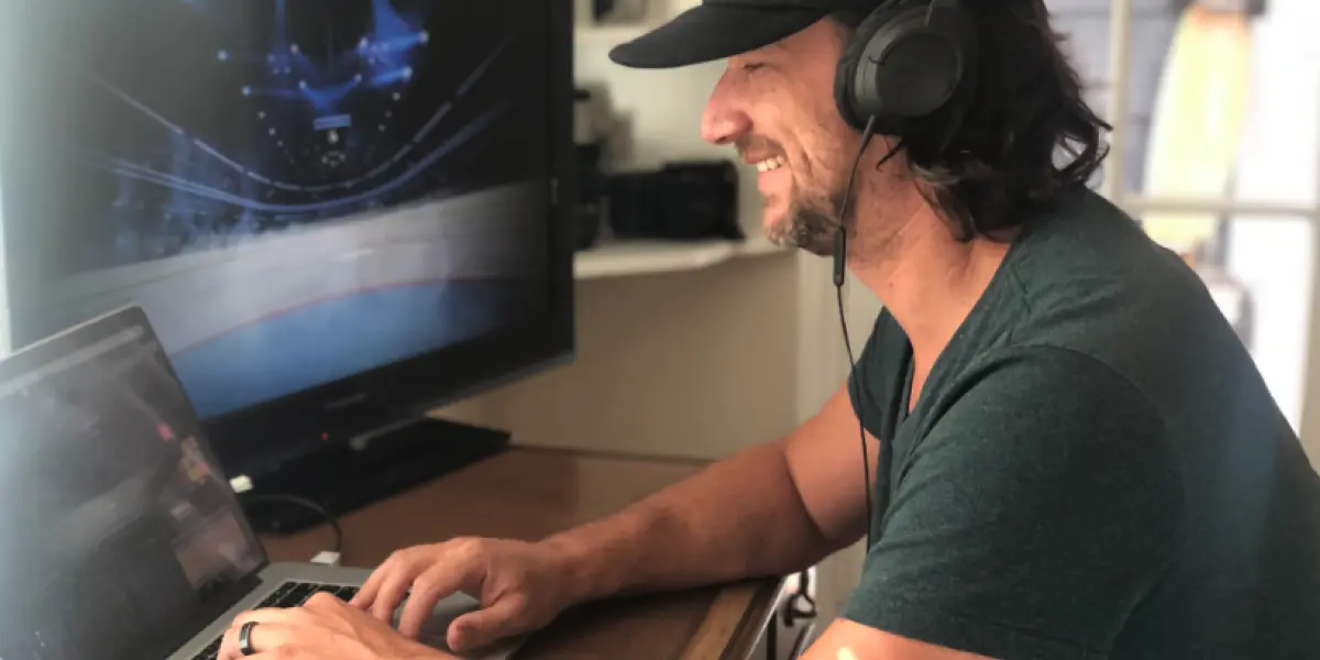 A man wearing headphones and a cap smiles while working on a laptop at a desk with a large screen displaying a sports scene.