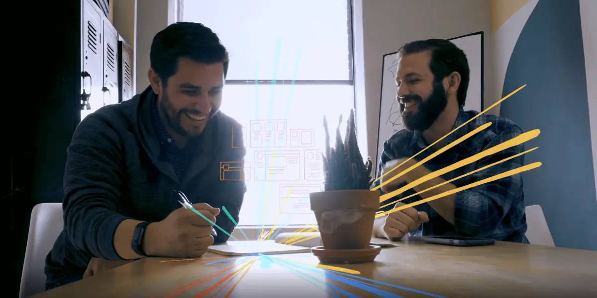 Two men brainstorm and laugh during a collaborative design session at a table with colorful visual elements added digitally.