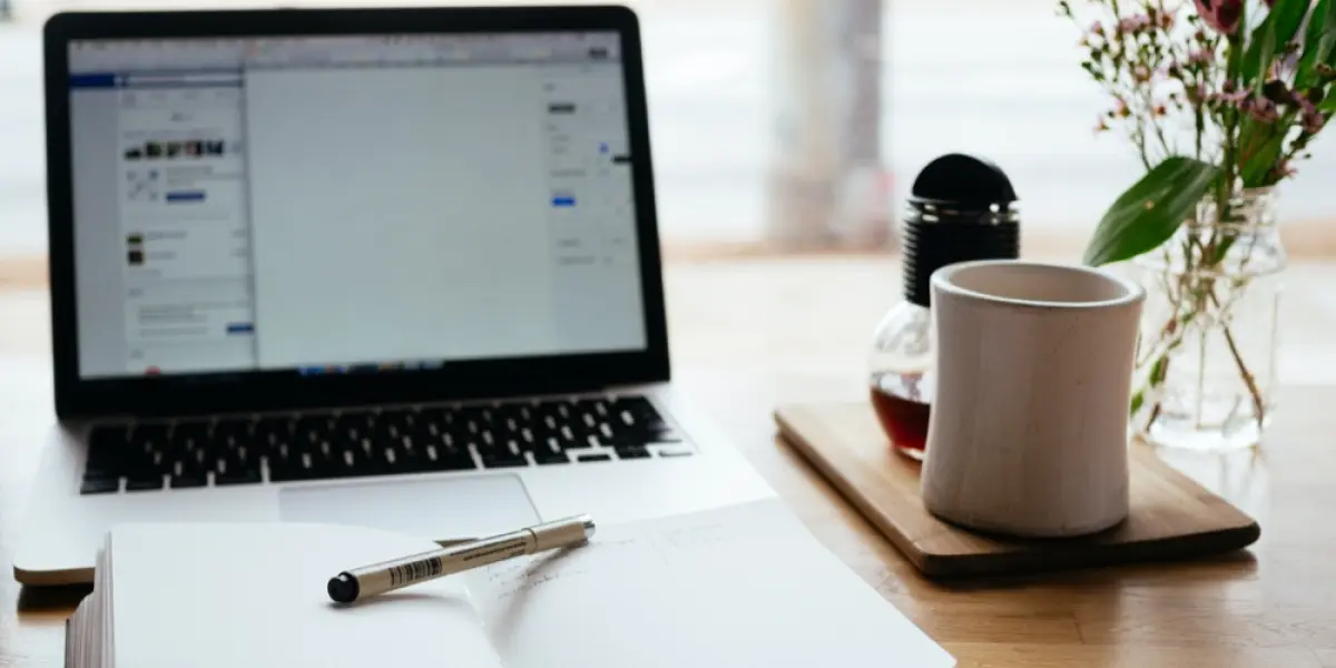 A cozy workspace features an open laptop, notebook with pen, and a coffee mug beside a jar of flowers.