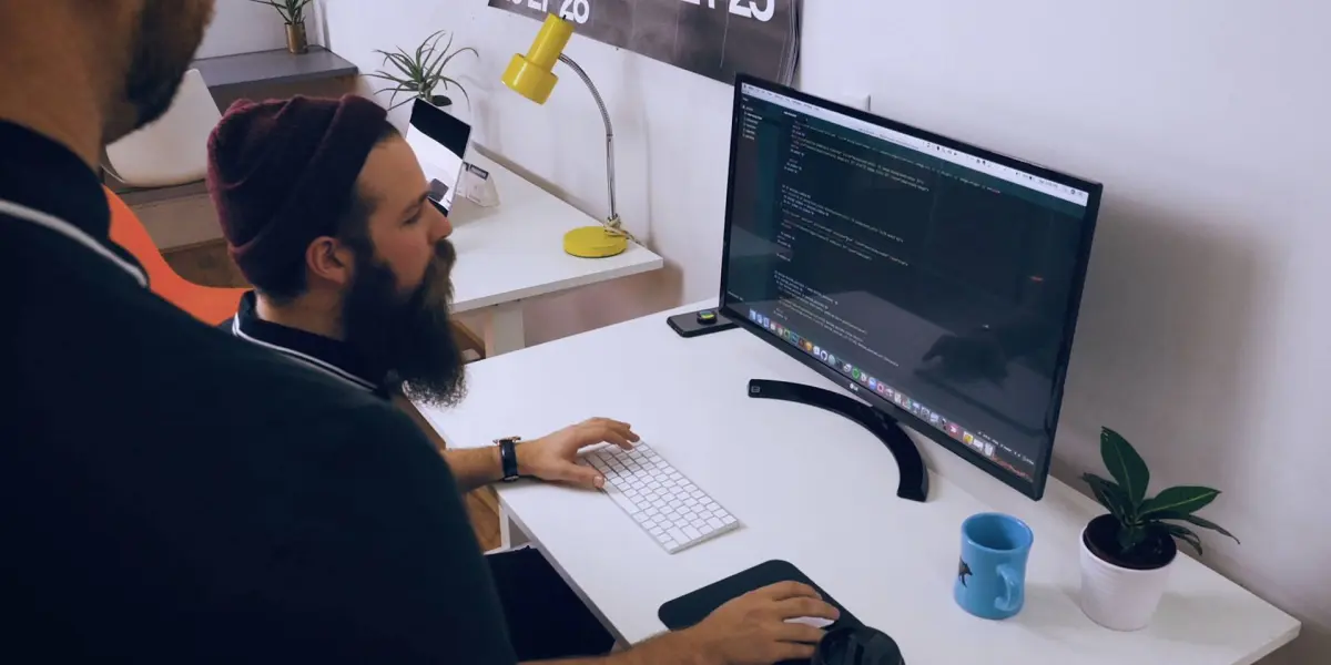 A bearded man works at a computer with code on a large monitor while another person observes beside him.
