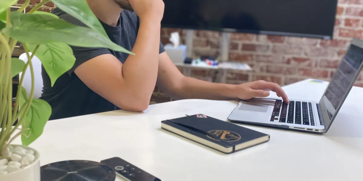 A man sits at a white desk focused on his laptop with a large monitor behind him and a notebook, remote, and plant nearby.