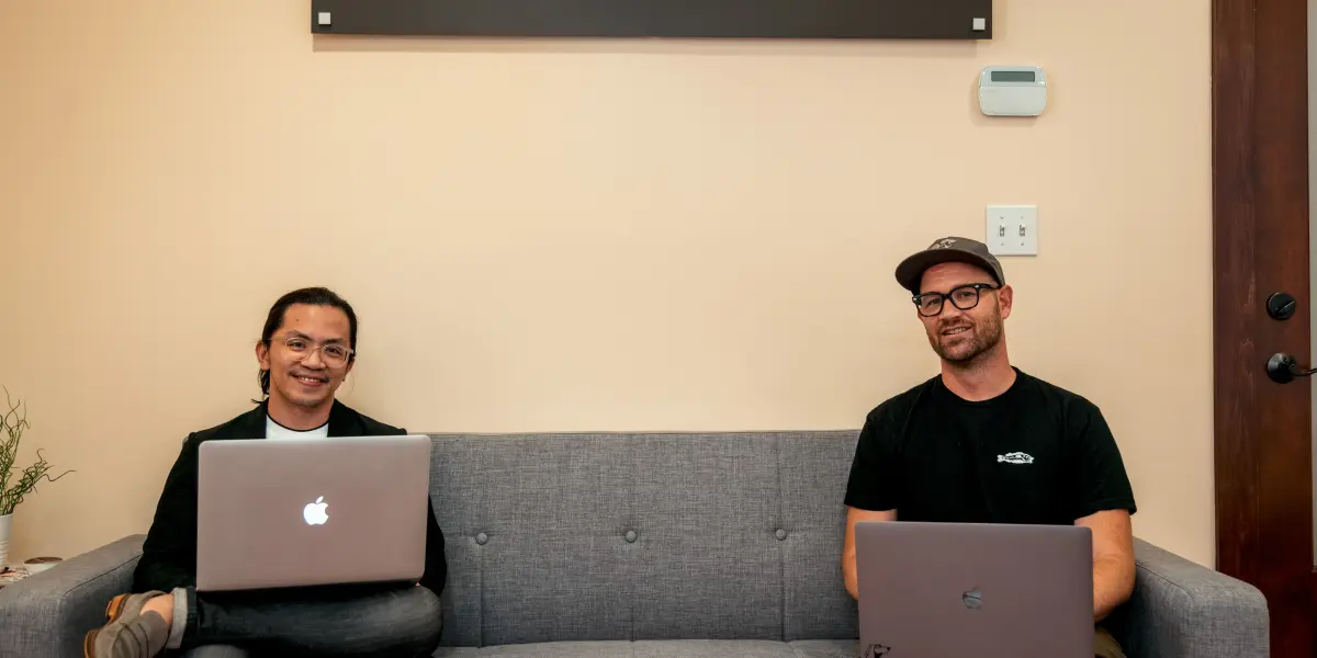 Two Hounder employees sit on a couch with laptops under a Hounder sign, smiling in a casual office setting.