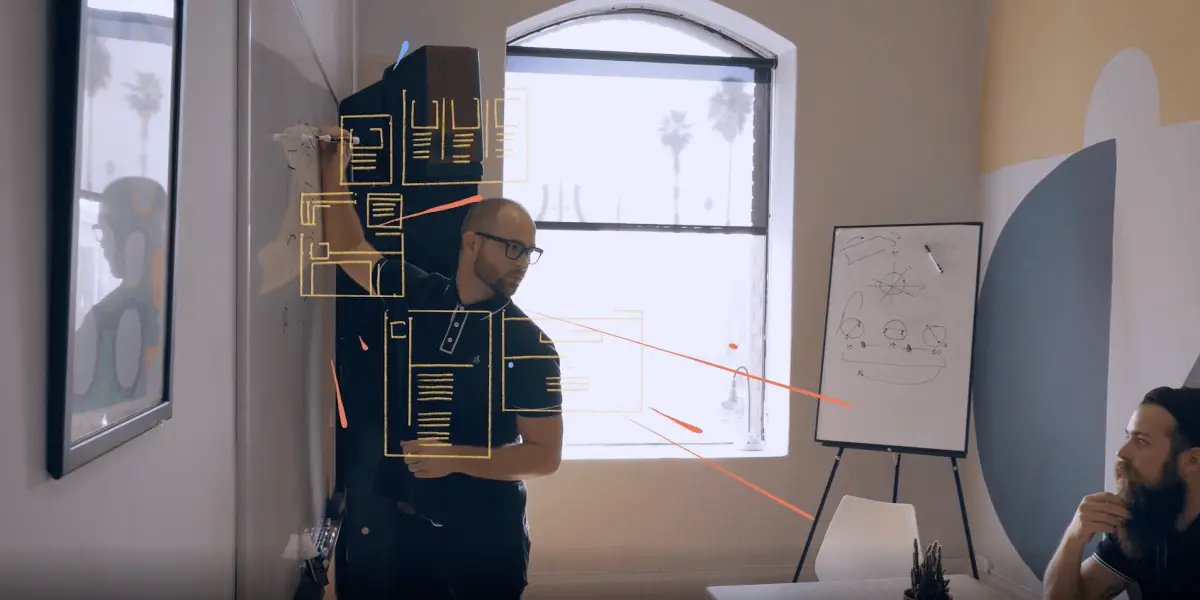 A presenter outlines a wireframe concept on a glass board while a teammate listens during a creative team meeting.