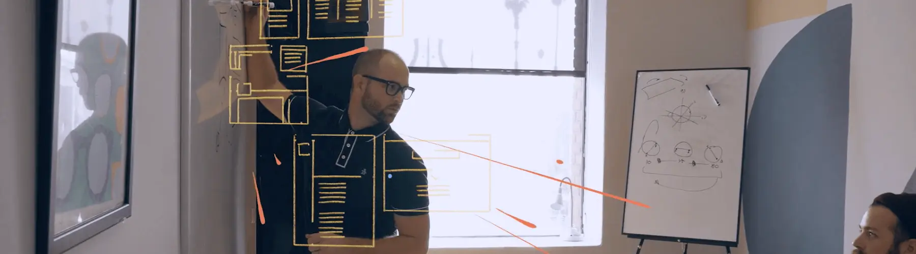 A presenter outlines a wireframe concept on a glass board while a teammate listens during a creative team meeting.