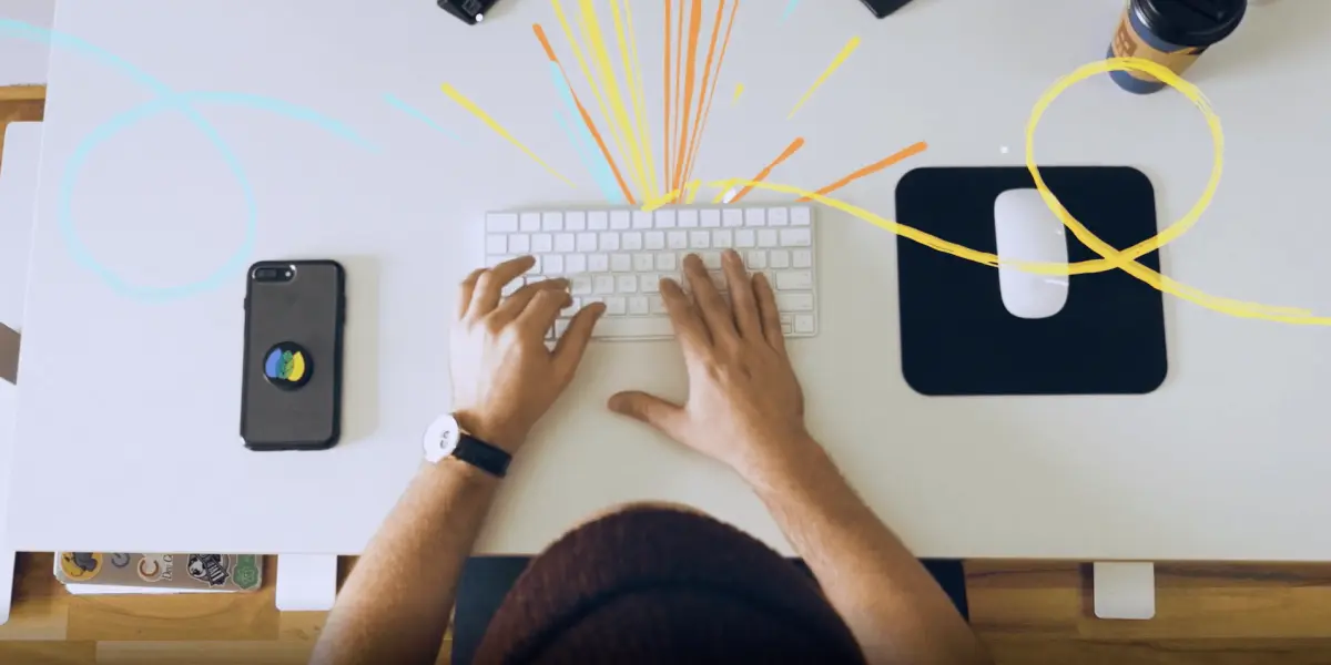 A Hounder employee types on a keyboard at a clean white desk, surrounded by creative tools and colorful design elements.