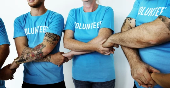 Volunteers wearing matching blue shirts stand in a circle, linking arms in a gesture of unity and teamwork.