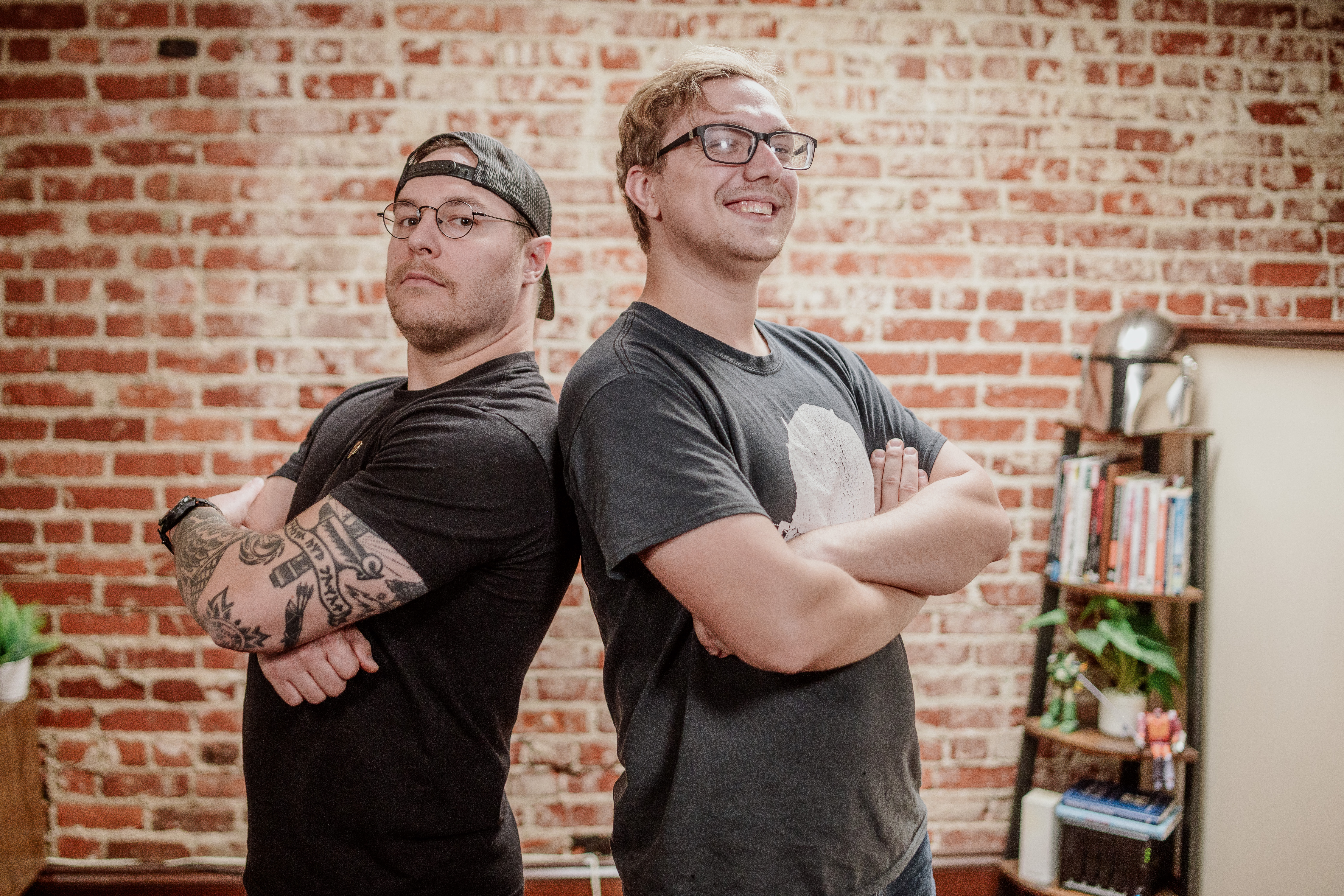 Two people standing back-to-back with arms crossed in front of a brick wall.