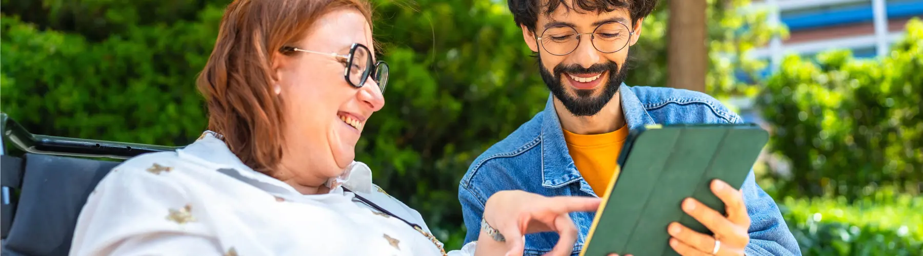 Two people smiling, looking at a tablet outdoors.