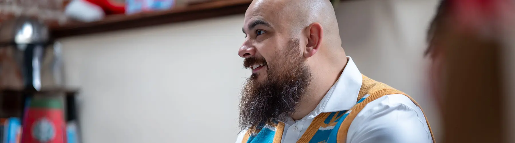 Man with a beard speaking indoors, wearing a colorful vest.