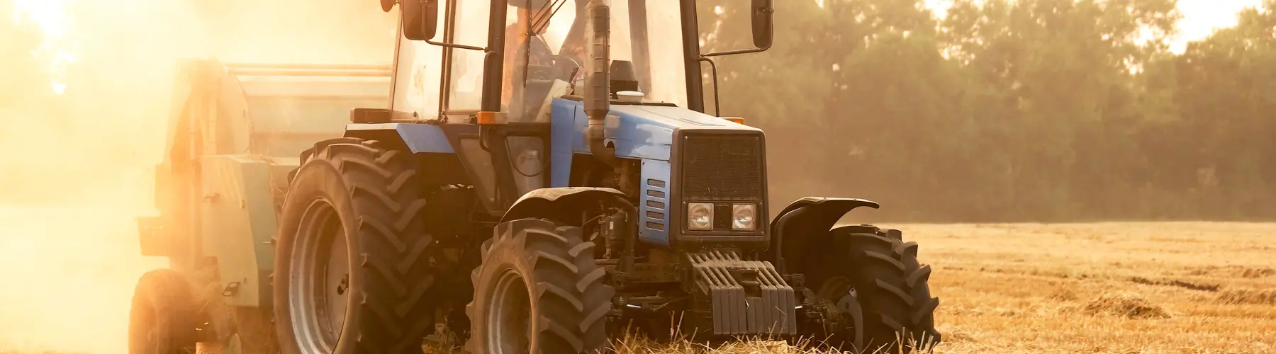 Blue tractor in a sunlit field, dust in the air, trees in the background.