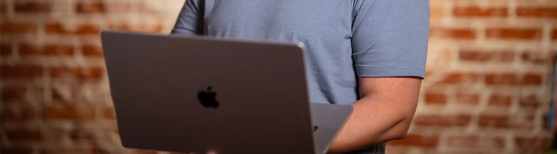 Person holding an open laptop against a brick wall backdrop.