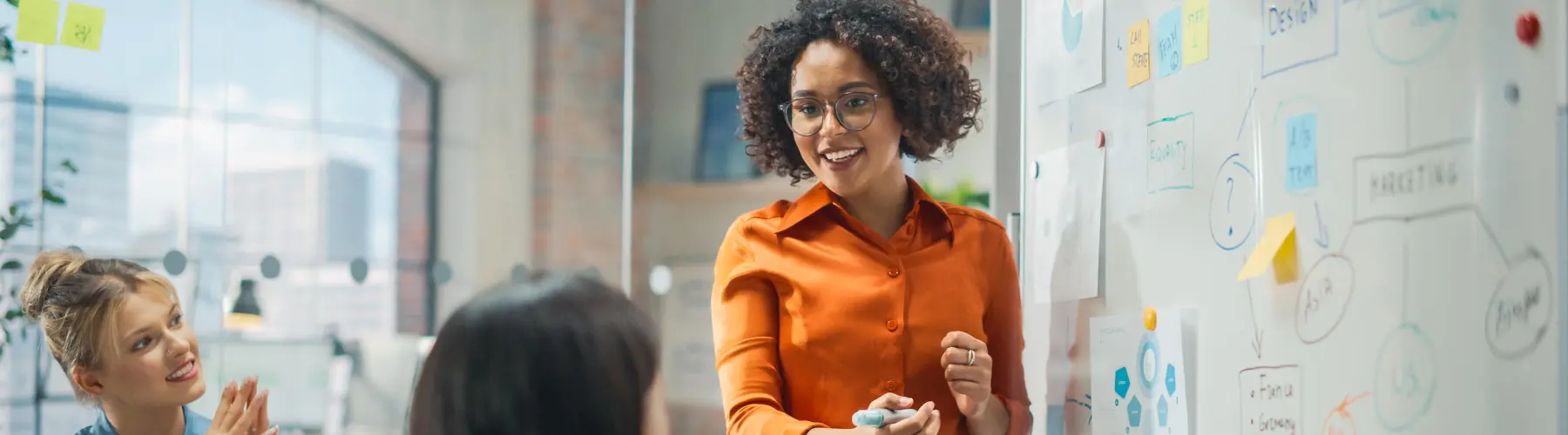 Woman smiling while presenting at a whiteboard in an office setting.