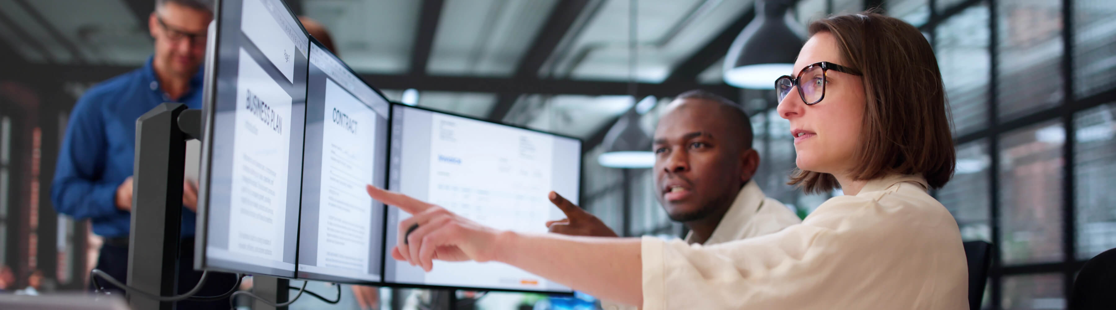 Two colleagues at computers in a modern office, discussing work.