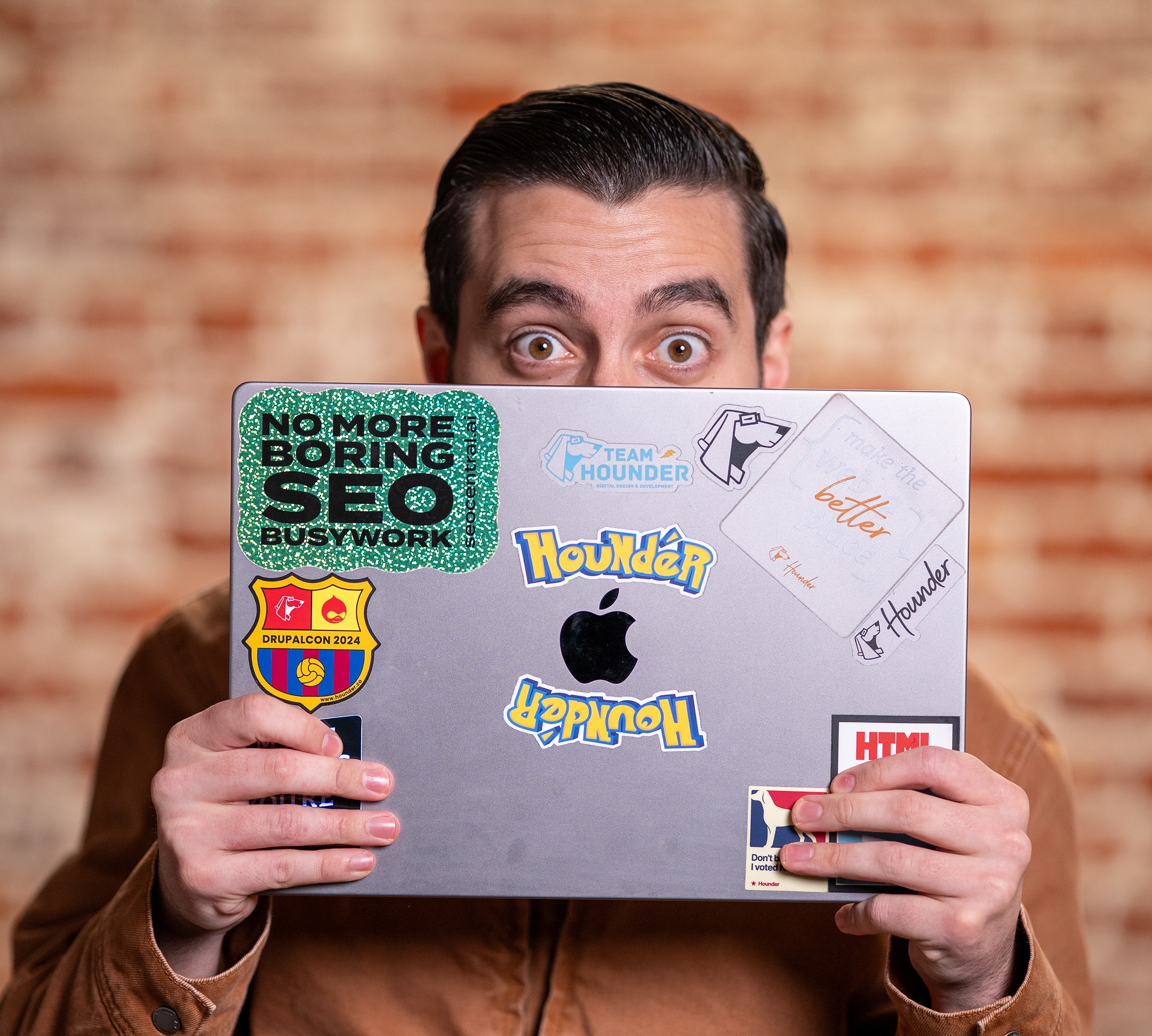 Man peeking over a laptop covered in colorful stickers against a brick wall.