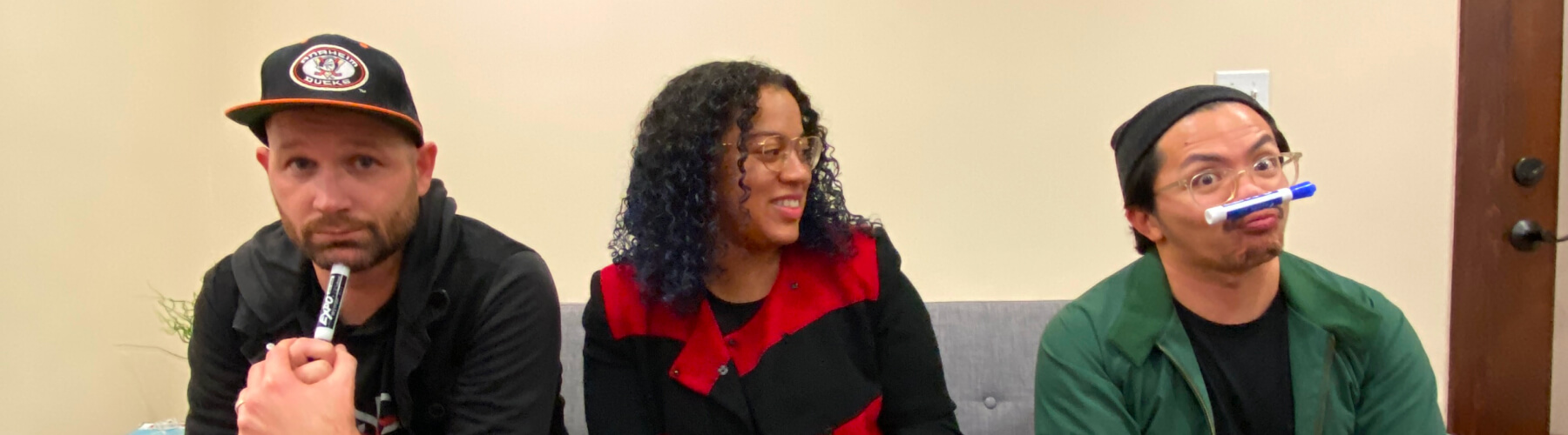 Three people sitting on a couch, two holding markers playfully.