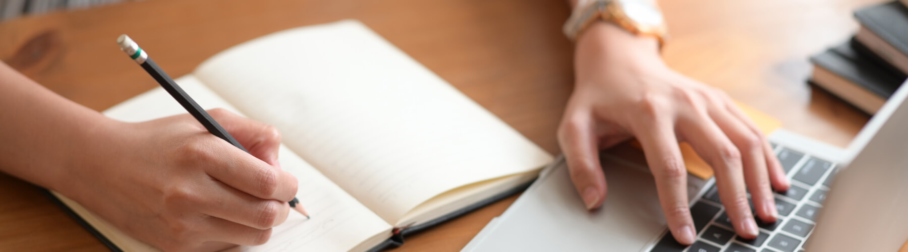 Person writing in a notebook while using a laptop on a desk.