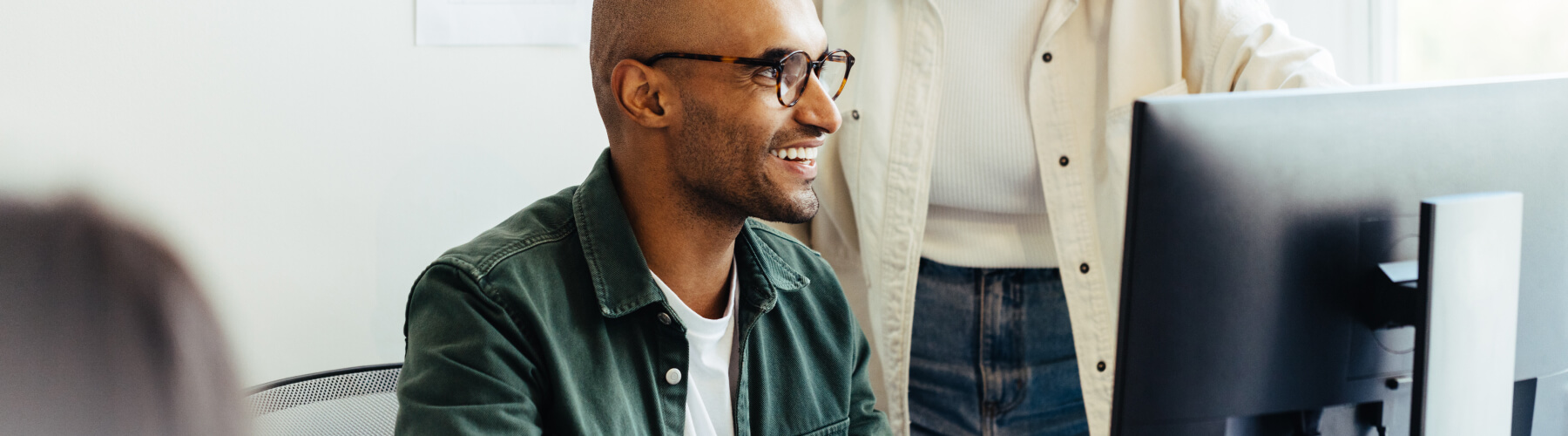 Smiling man with glasses working on a computer with a colleague nearby.