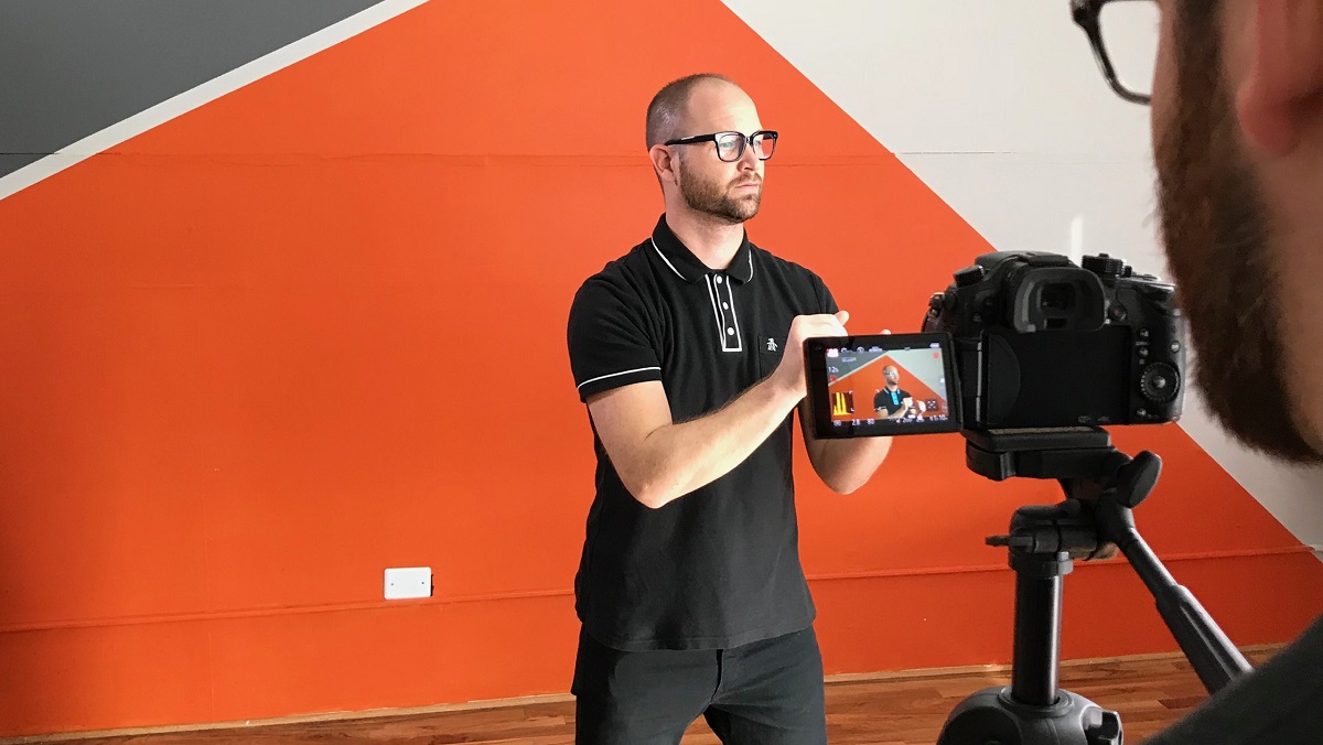 Man being filmed with a camera against an orange geometric wall.