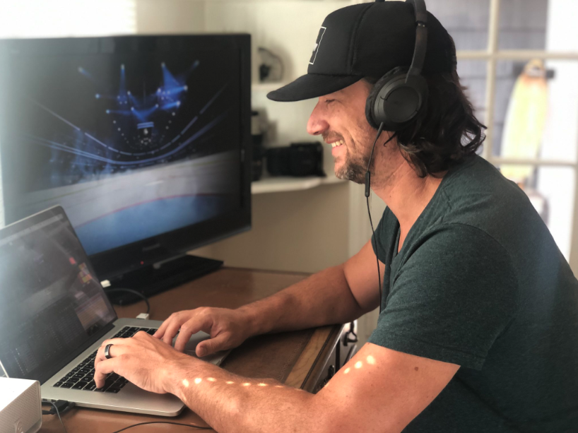 Smiling man with headphones working on a laptop beside a monitor in a cozy room.