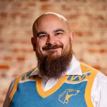 Smiling man in a blue and gold sweater, brick wall background.