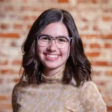 Smiling woman with glasses, brown hair, wearing a floral top against a brick wall background.