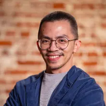 Smiling person with glasses and a blue jacket against a brick wall.