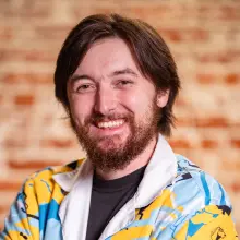 Smiling man with a beard in a colorful jacket against a blurred brick wall.