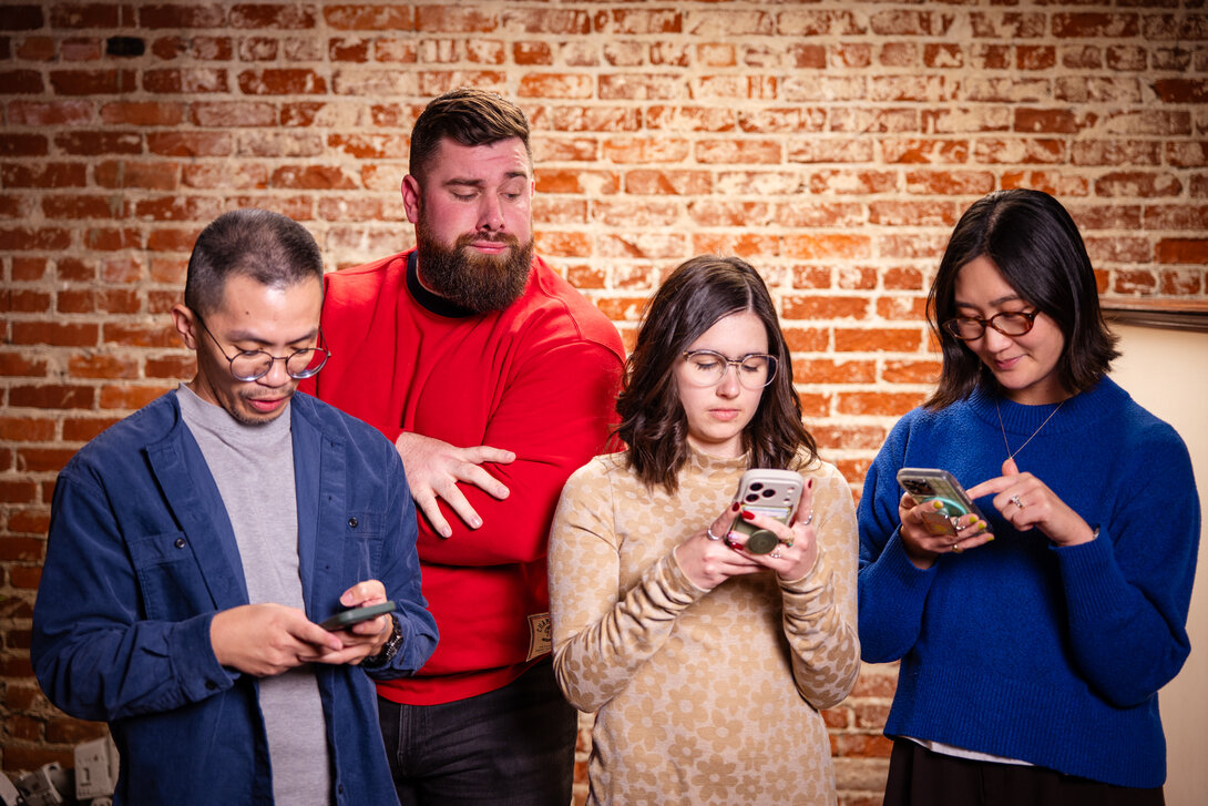 Four people standing, using smartphones, smiling, against a brick wall.