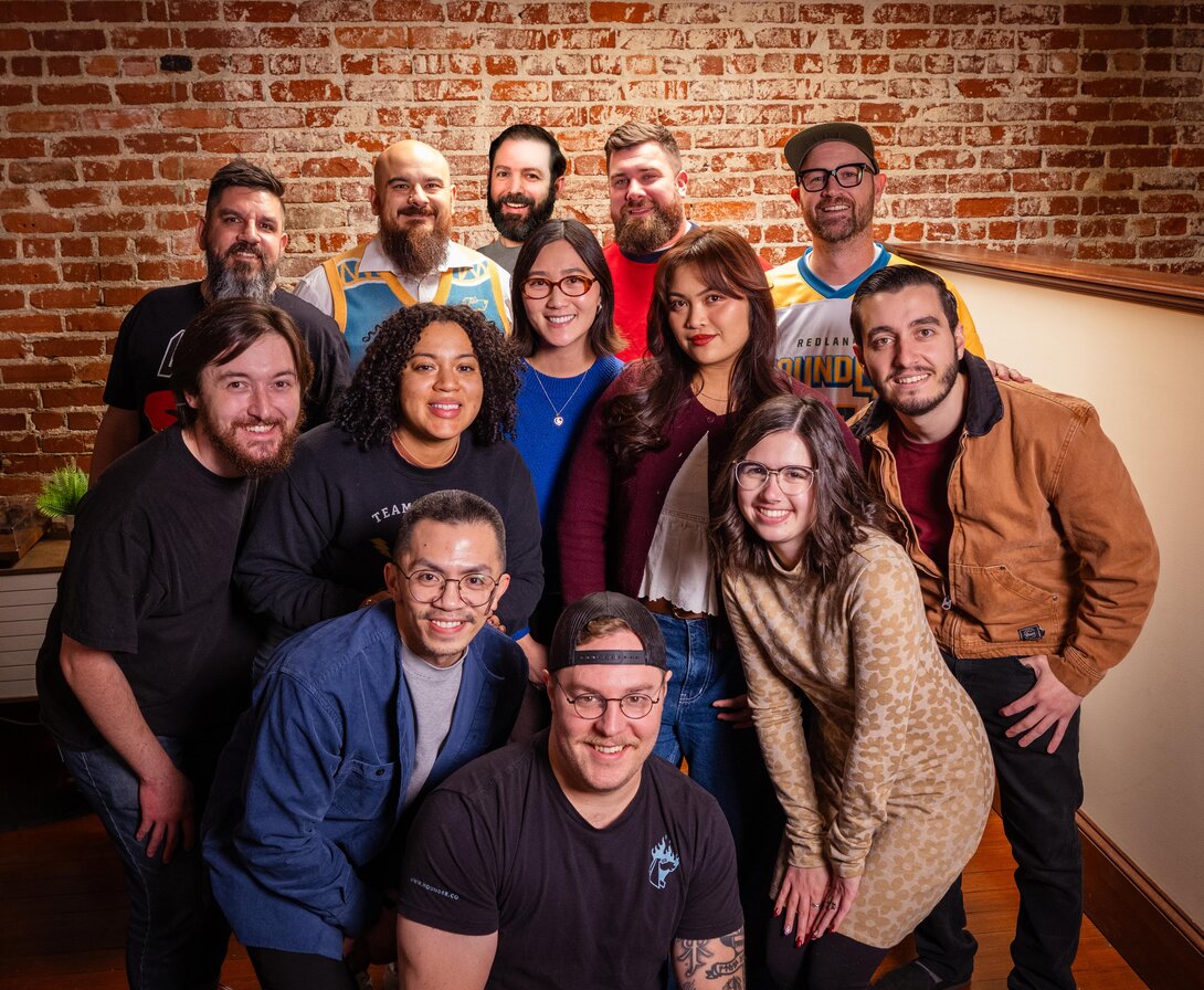 The Hounder team poses together for a group photo in front of a brick wall, smiling and dressed casually in a collaborative office setting.