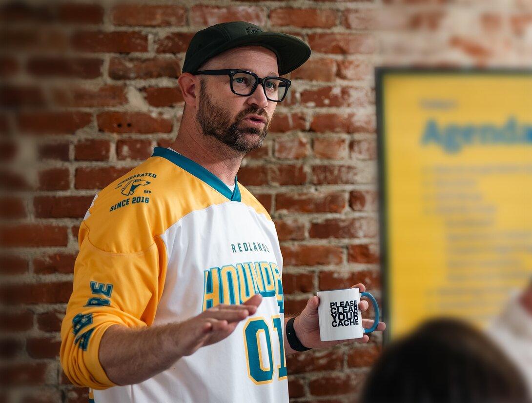 Man in jersey and cap speaking, holding a coffee mug, with brick wall background.