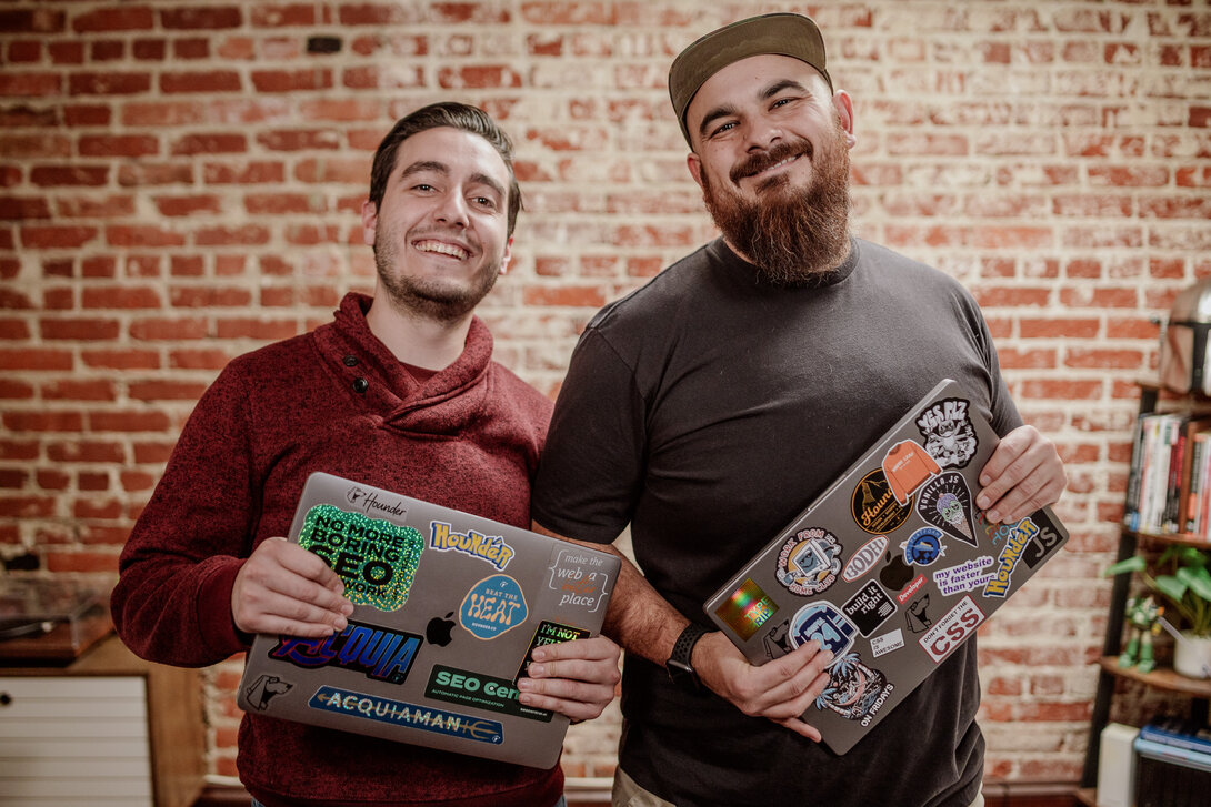 Two smiling men holding laptops with colorful stickers against a brick wall.