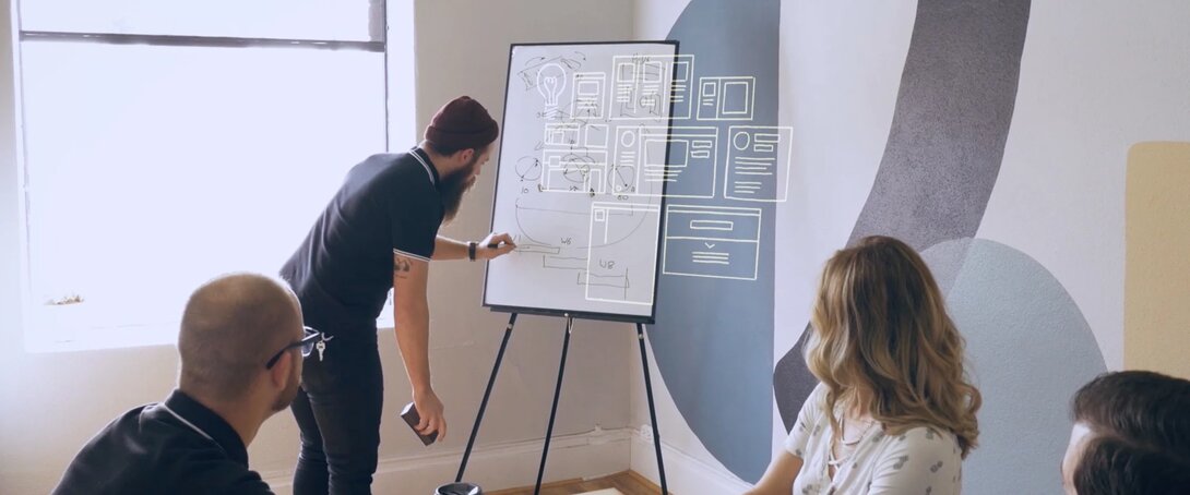 Team brainstorming session with a man writing on a whiteboard in a creative office setting.