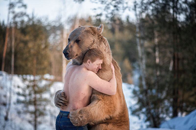 Shirtless man hugging a large brown bear in a snowy forest.