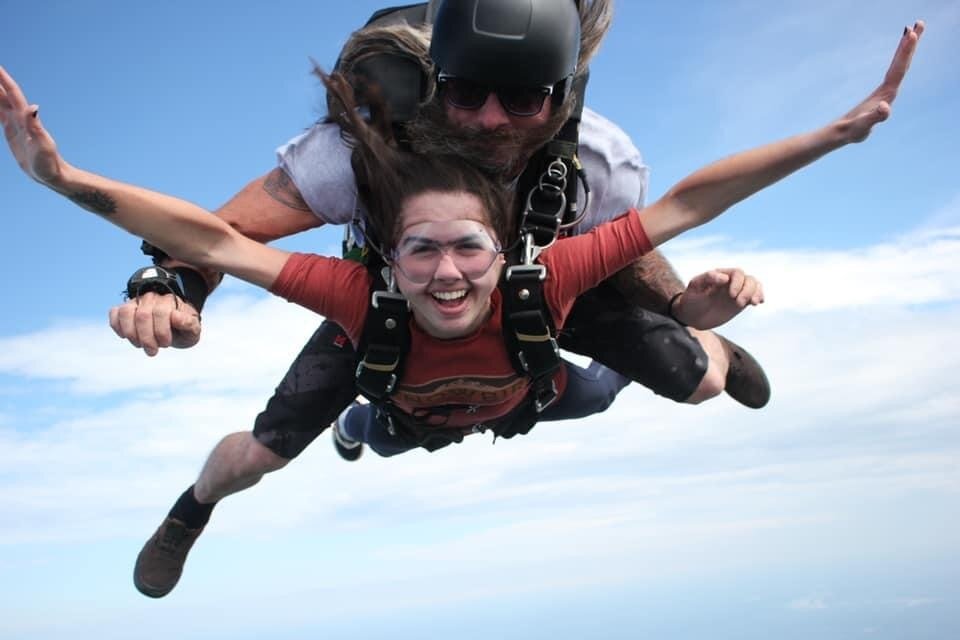 Tandem skydivers freefalling with smiles against a clear blue sky.