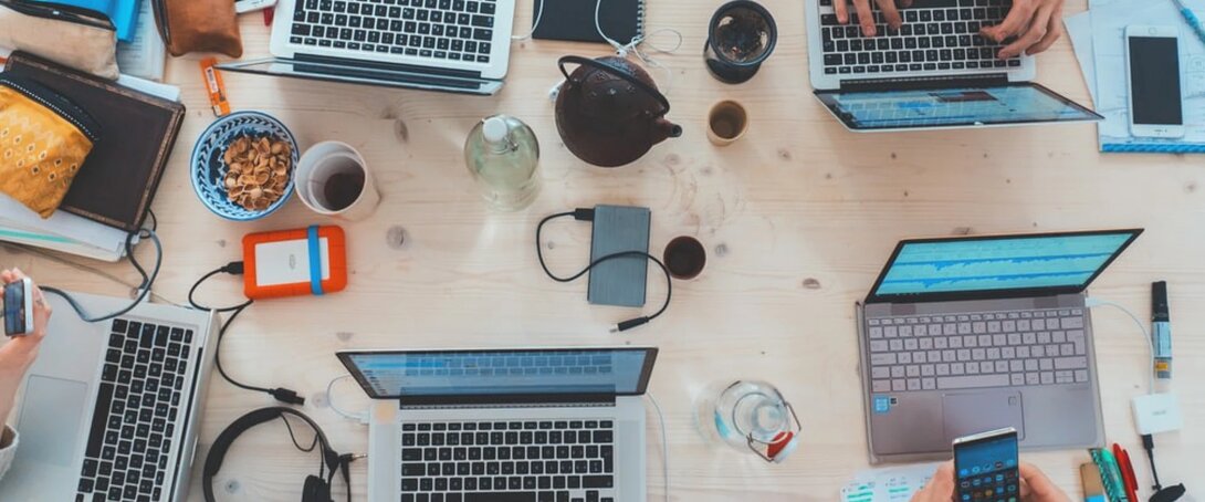 Overhead view of laptops and gadgets on a wooden desk, with people working.