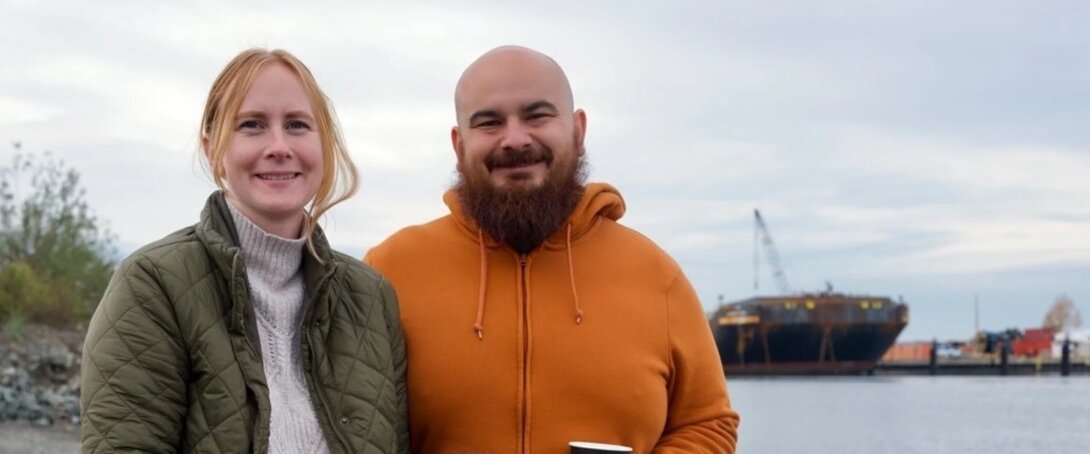 Man and woman smiling near waterfront with ship in background.