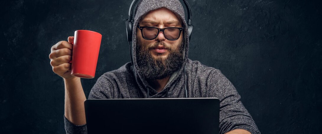 Bearded man with headphones and a red mug working on a laptop.