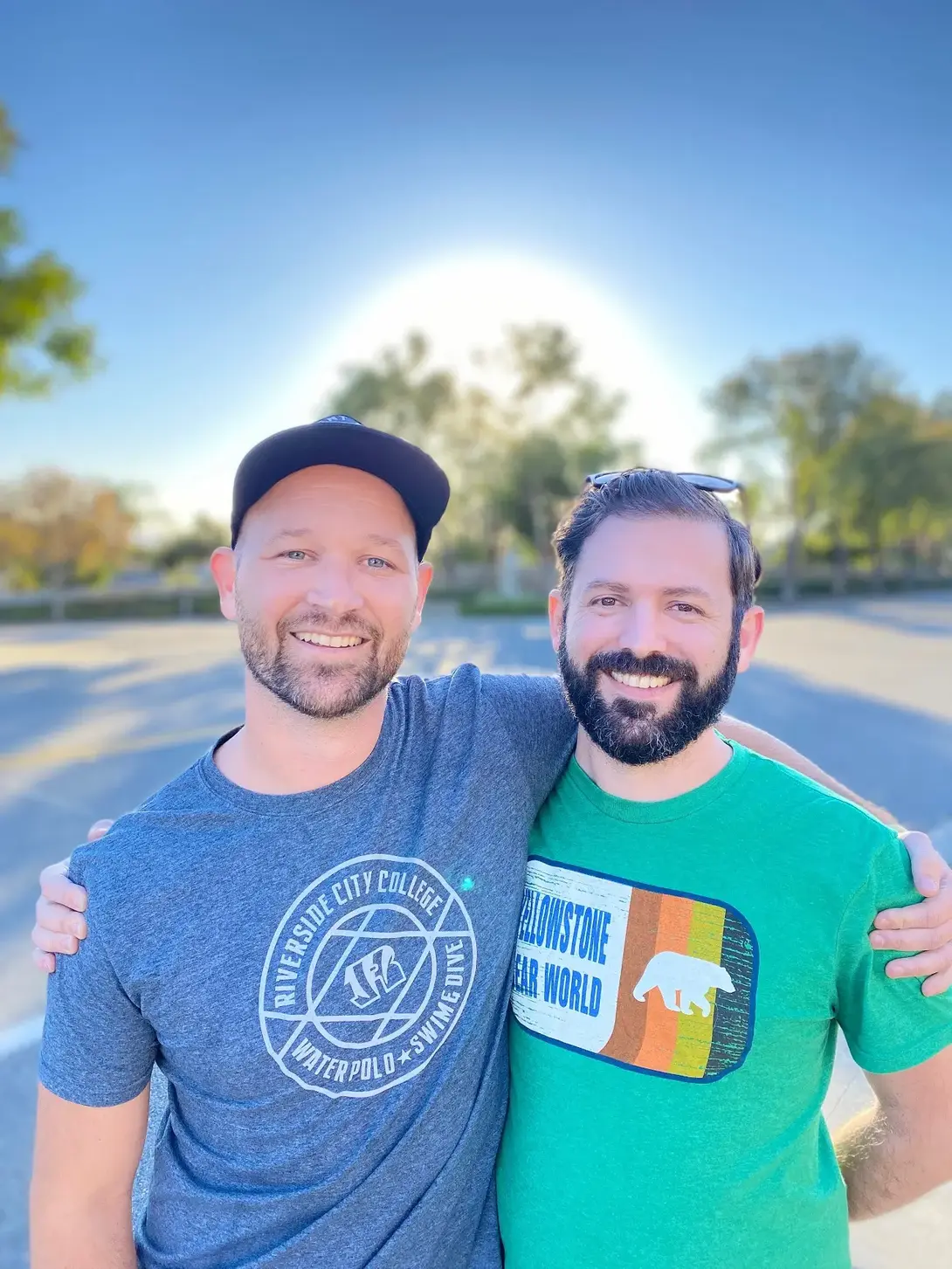 Two friends smiling in casual t-shirts outdoors, with trees and a clear sky in the background.
